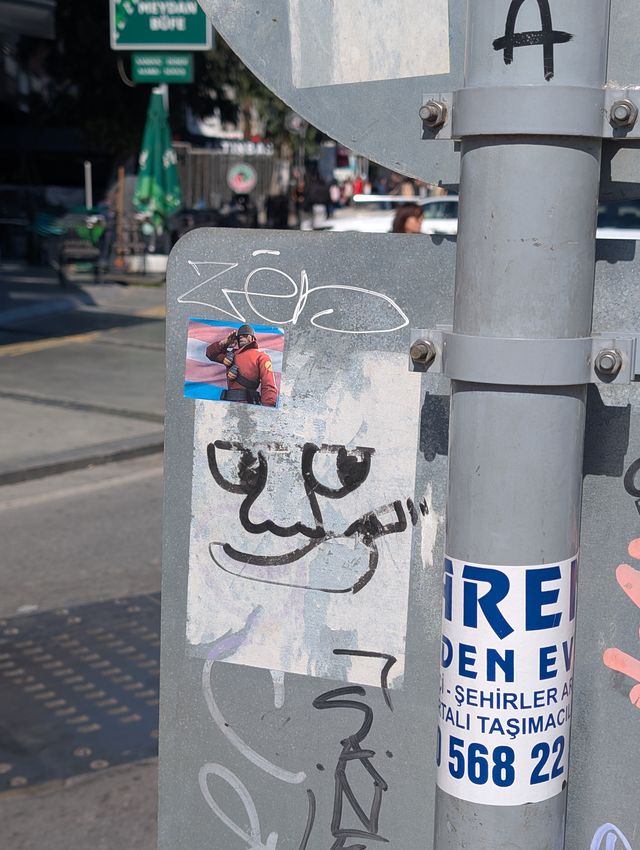 A grey metal street pole covered in graffiti and stickers. Near the top, a small sticker depicts the Soldier from Team Fortress 2 saluting in front of a transgender pride flag. Below it, a large minimalistic hand-drawn graffiti face with round hollow eyes and a cigarette hanging from the corner of its mouth. A busy Turkish street with café umbrellas is visible in the blurred background.