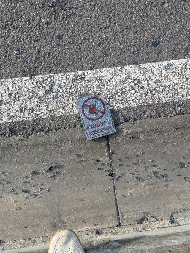 A broken piece of a sign lying on asphalt among shattered glass, displaying a "no market trolleys" symbol and Turkish text.